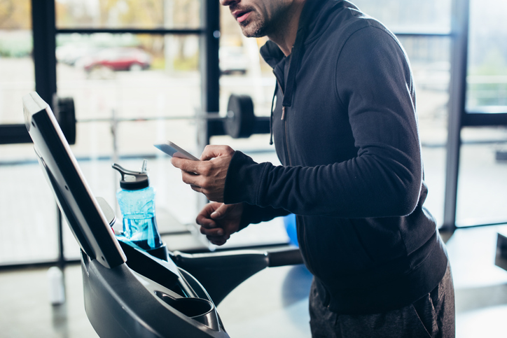 Man in blue hoodie looking at a cell phone instead of focusing on his workout.